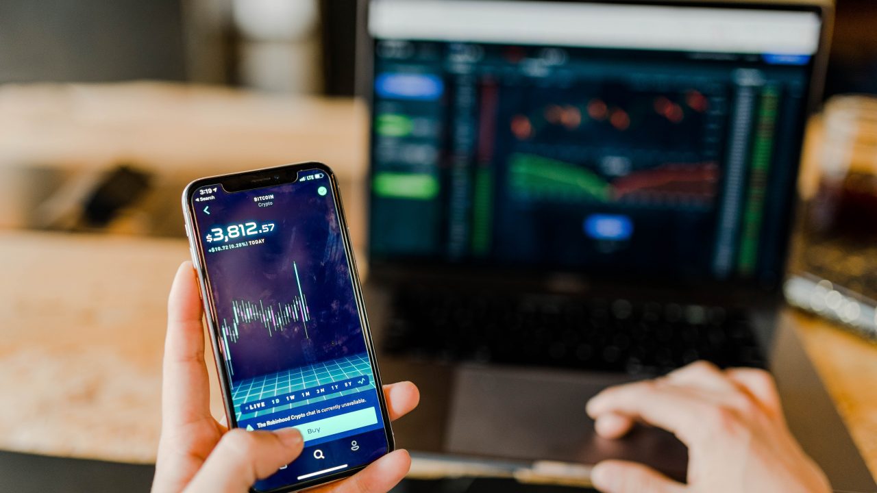someone holding up their phone showing a graph of increasing returns while typing on a computer with the other hand