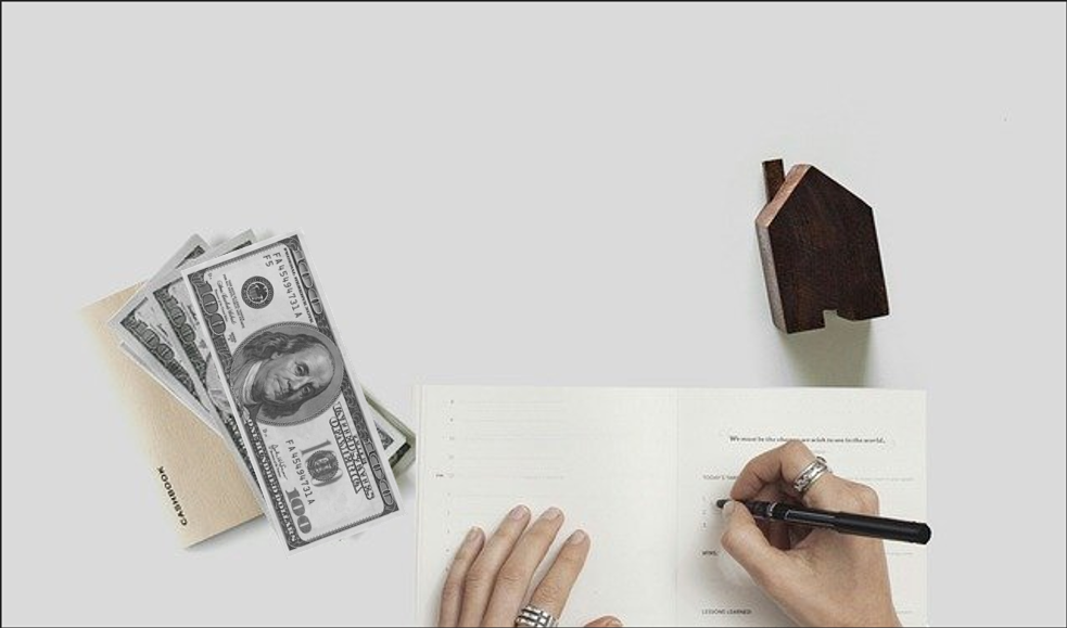 someone's hands filling out a tax form next to a wooden house ornament and a few hundred dollar bills