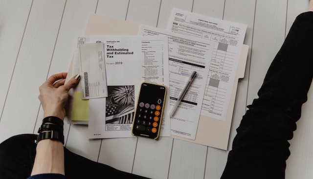 a hand holding a check beside tax forms and a calculator open on their phone
