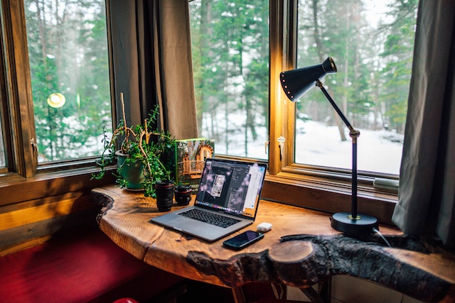 a home office with lamp, computer, and phone on a wood desk with a window overlooking snow covered forest