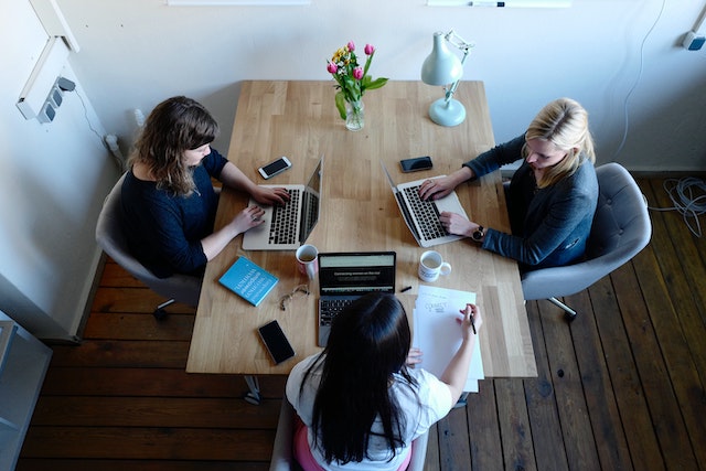 a bird’s eye view of three businesswomen seated at a shared table working on laptops