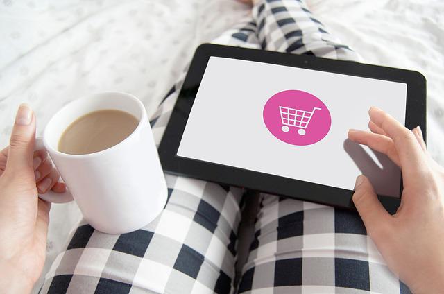 a tablet with a shopping cart on someone's legs wearing pajamas and a coffee in their left hand