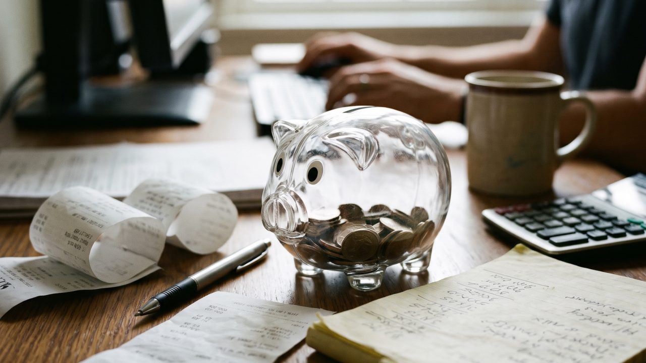 a cluttered desk with a transparent piggy bank, some receipts and loose stationery on the surface
